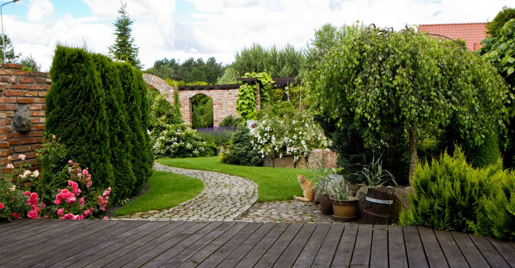 Aménagement jardin paysager avec allée pavée et fleurs Jardin paysager avec allée en pavés, roses roses, chat roux et terrasse en bois sous un ciel nuageux.