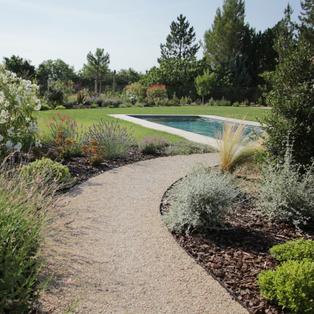Jardin paysager avec piscine, allée en gravier sinueuse, lavande et massifs fleuris sous un ciel bleu.