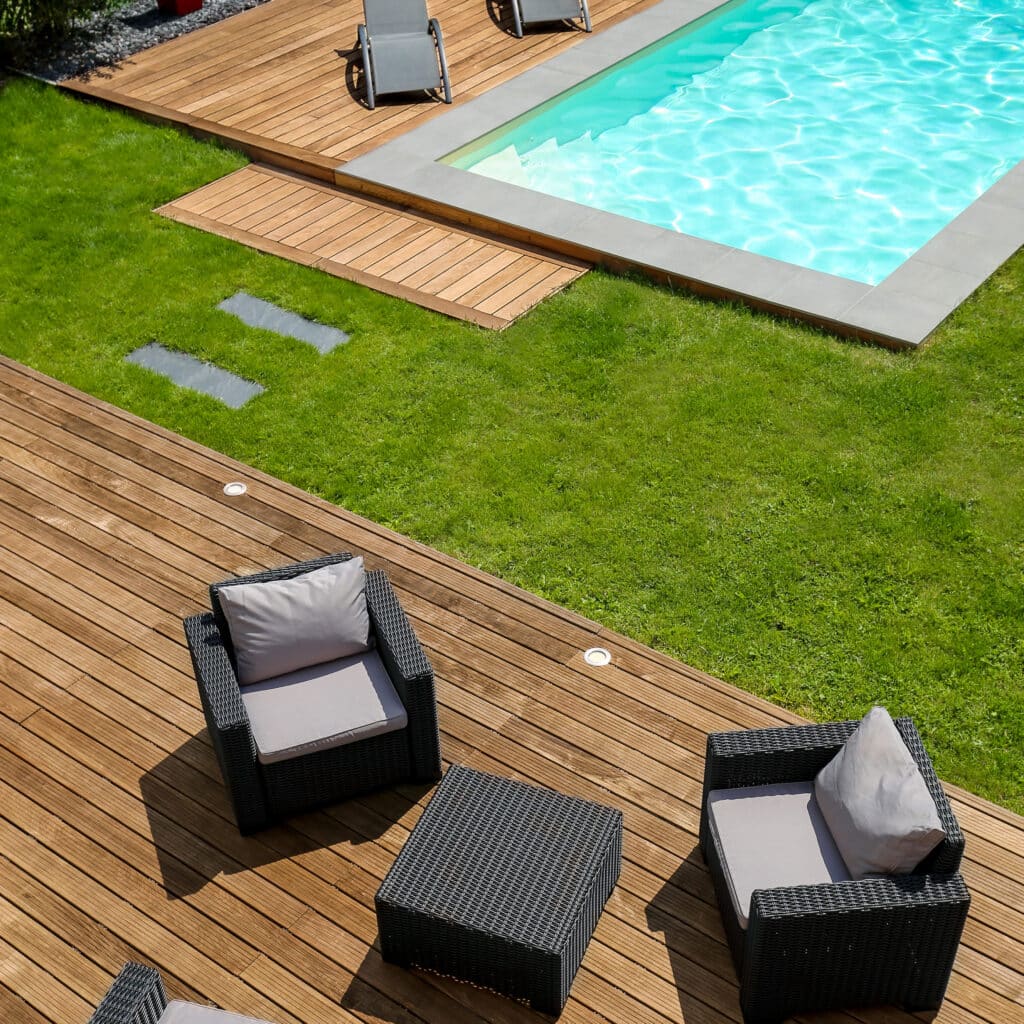 Aménagement de terrasse bois moderne avec piscine et jardin Terrasse en bois avec fauteuils noirs et table basse près d'une piscine turquoise et d'une pelouse verte.